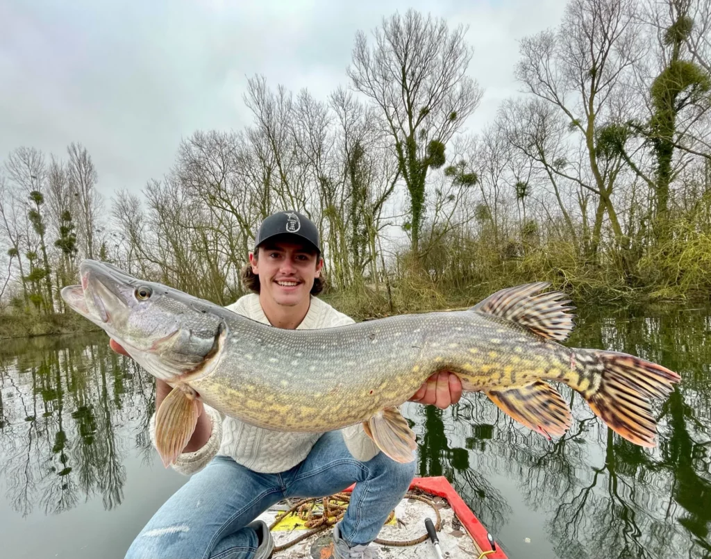Un pêcheur en pull clair présente un brochet au-dessus d’une barque.