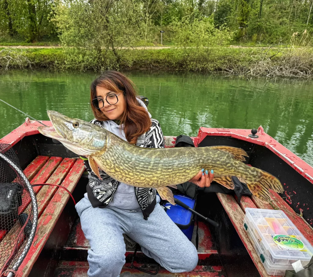 Une pêcheuse présente un brochet à bord d’une barque rouge.