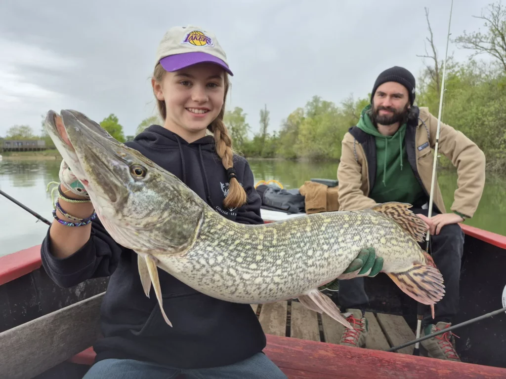 Une jeune pêcheuse présente un brochet, accompagnée d’un adulte dans la barque.