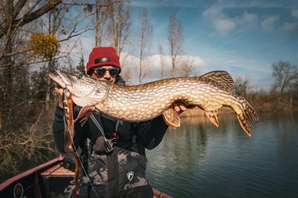Un pêcheur en bonnet rouge présente un brochet avec un leurre souple orange.